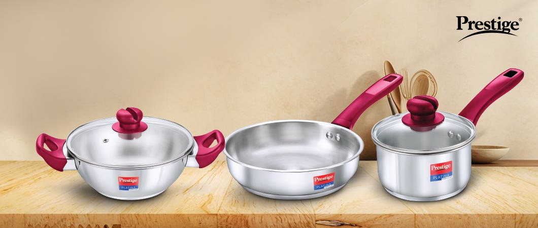 Prestige stainless-steel cookware set, including a frying pan and a saucepan with red handles, displayed with kitchen utensils.
